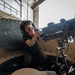 A young child wearing ear protection headphones and a green wristband is seated in the turret hatch of a military armored vehicle, gripping a mounted machine gun. The setting appears to be an indoor military hangar or exhibition facility, suggesting this is a public open day or air show event. The child is smiling, indicating this is a supervised interactive display rather than an operational context.