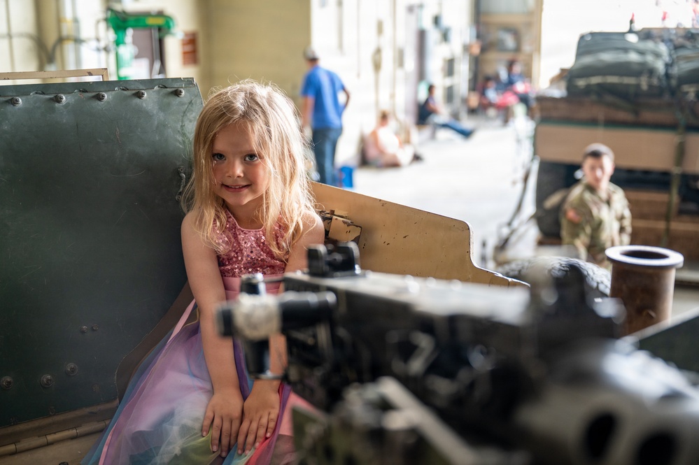 A young blonde girl wearing a pink sequined and rainbow tulle dress is seated inside or beside a large military vehicle, likely at a military museum or open day event. She is smiling at the camera while surrounded by heavy metal machinery and equipment. In the background, other visitors including a boy in camouflage clothing and adults can be seen exploring the facility.