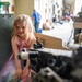 A young blonde girl wearing a pink sequined and rainbow tulle dress is seated inside or beside a large military vehicle, likely at a military museum or open day event. She is smiling at the camera while surrounded by heavy metal machinery and equipment. In the background, other visitors including a boy in camouflage clothing and adults can be seen exploring the facility.
