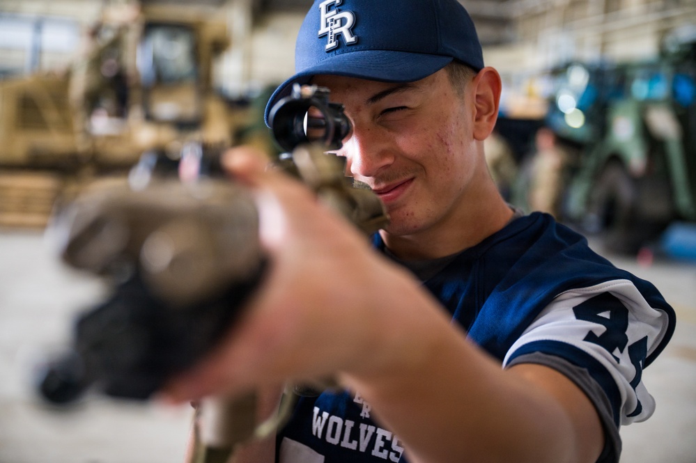 A young person wearing a blue 'FR Wolves' baseball cap and jersey is looking through the scope of a firearm, smiling. The setting appears to be a military hangar or maintenance facility, with large vehicles visible in the blurred background. This appears to be a demonstration or familiarization event, likely a military outreach or youth program.