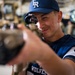 A young person wearing a blue 'FR Wolves' baseball cap and jersey is looking through the scope of a firearm, smiling. The setting appears to be a military hangar or maintenance facility, with large vehicles visible in the blurred background. This appears to be a demonstration or familiarization event, likely a military outreach or youth program.