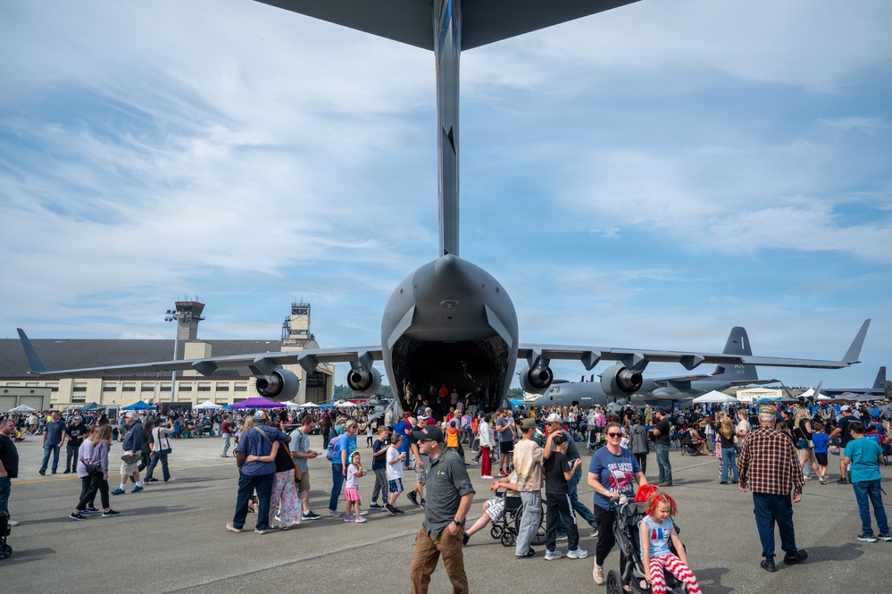 A large military airshow or open house event at a military airbase, with crowds of civilians gathered around a large military transport aircraft, likely a C-17 Globemaster III, viewed from directly beneath its tail section. The aircraft's rear cargo ramp appears open, allowing visitors to walk inside. A control tower and large hangars are visible in the background.