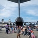 A large military airshow or open house event at a military airbase, with crowds of civilians gathered around a large military transport aircraft, likely a C-17 Globemaster III, viewed from directly beneath its tail section. The aircraft's rear cargo ramp appears open, allowing visitors to walk inside. A control tower and large hangars are visible in the background.