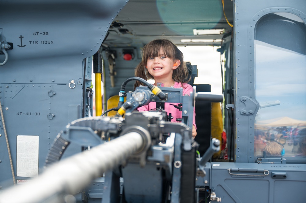 A young girl wearing a pink shirt is smiling inside the cabin of a military helicopter, likely during a public airshow or open house event. She is standing near a mounted weapon system (machine gun) in the open door of the aircraft. The setting appears to be an outdoor aviation display where the public is allowed to explore military equipment.