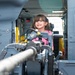 A young girl wearing a pink shirt is smiling inside the cabin of a military helicopter, likely during a public airshow or open house event. She is standing near a mounted weapon system (machine gun) in the open door of the aircraft. The setting appears to be an outdoor aviation display where the public is allowed to explore military equipment.