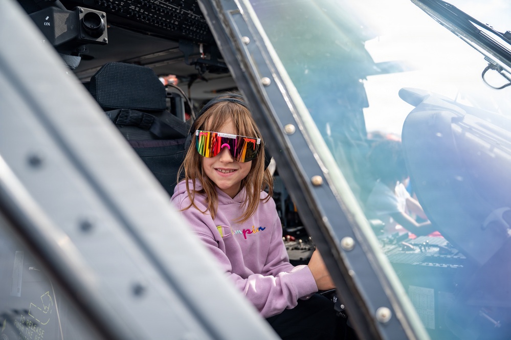 A young girl is seated inside the cockpit of a military or large aircraft, smiling at the camera. She is wearing colorful mirrored wraparound sunglasses and a pink hoodie with the word 'Dumplin' written on it. This appears to be a public airshow or static display event where visitors are allowed to sit in aircraft.