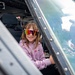 A young girl is seated inside the cockpit of a military or large aircraft, smiling at the camera. She is wearing colorful mirrored wraparound sunglasses and a pink hoodie with the word 'Dumplin' written on it. This appears to be a public airshow or static display event where visitors are allowed to sit in aircraft.