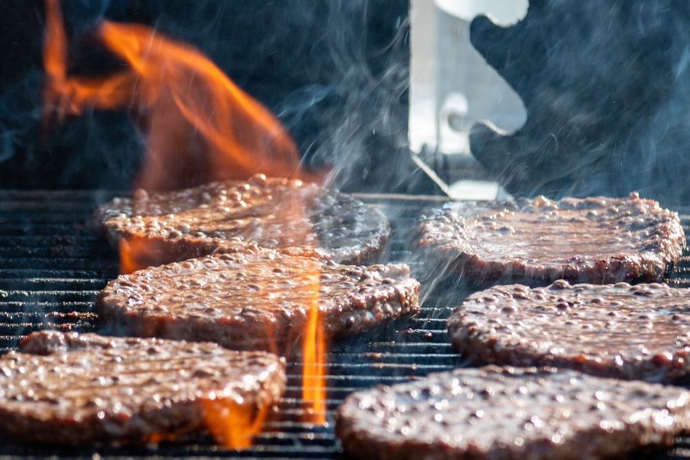 Multiple beef burger patties are being grilled on a metal grate over an open flame. The patties are actively cooking with visible flames, steam, and sizzling juices. The close-up shot captures the intense heat and cooking process in vivid detail.