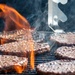 Multiple beef burger patties are being grilled on a metal grate over an open flame. The patties are actively cooking with visible flames, steam, and sizzling juices. The close-up shot captures the intense heat and cooking process in vivid detail.
