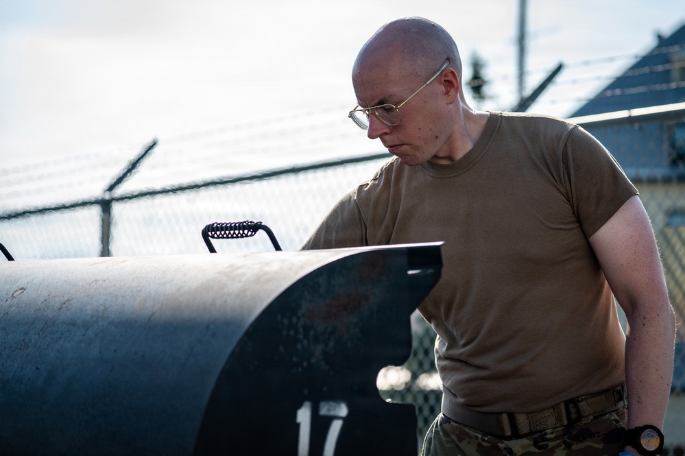A bald individual wearing military-style olive drab clothing and wire-rimmed glasses is leaning over and inspecting a large dark cylindrical object marked with the number '17'. The setting appears to be a secured outdoor area with chain-link fencing topped with razor wire visible in the background. The image has a professional, documentary-style composition with strong directional lighting.