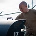 A bald individual wearing military-style olive drab clothing and wire-rimmed glasses is leaning over and inspecting a large dark cylindrical object marked with the number '17'. The setting appears to be a secured outdoor area with chain-link fencing topped with razor wire visible in the background. The image has a professional, documentary-style composition with strong directional lighting.
