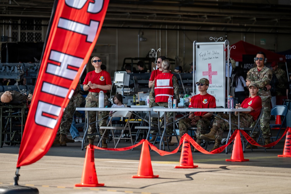 Military personnel are staffing a First Aid Station at what appears to be an outdoor airshow or military open house event. Several service members in red shirts and camouflage uniforms are positioned behind a table near a whiteboard marked with a red cross. The area is cordoned off with orange safety cones and red caution tape.