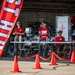 Military personnel are staffing a First Aid Station at what appears to be an outdoor airshow or military open house event. Several service members in red shirts and camouflage uniforms are positioned behind a table near a whiteboard marked with a red cross. The area is cordoned off with orange safety cones and red caution tape.
