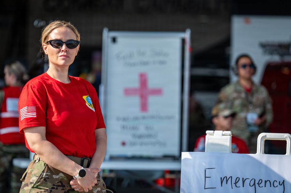 A military medical personnel member wearing a red t-shirt with a US Air Force patch and American flag sleeve patch stands at what appears to be a medical aid station or triage area. She is wearing camouflage pants, sunglasses, and a watch, with her hands clasped in front of her. Behind her are Red Cross signage, an 'Emergency' sign, medical equipment, and other personnel in military uniforms.