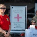 A military medical personnel member wearing a red t-shirt with a US Air Force patch and American flag sleeve patch stands at what appears to be a medical aid station or triage area. She is wearing camouflage pants, sunglasses, and a watch, with her hands clasped in front of her. Behind her are Red Cross signage, an 'Emergency' sign, medical equipment, and other personnel in military uniforms.