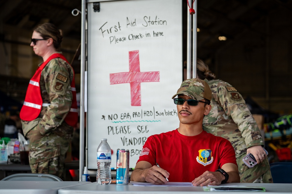 A military first aid station is set up, likely at an outdoor event or airshow. A service member in a red t-shirt and camouflage cap sits at a check-in table in the foreground, while two other personnel in OCP (Operational Camouflage Pattern) uniforms are visible in the background. A whiteboard sign clearly identifies the location as a 'First Aid Station' with check-in instructions and notes about missing items.
