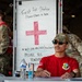 A military first aid station is set up, likely at an outdoor event or airshow. A service member in a red t-shirt and camouflage cap sits at a check-in table in the foreground, while two other personnel in OCP (Operational Camouflage Pattern) uniforms are visible in the background. A whiteboard sign clearly identifies the location as a 'First Aid Station' with check-in instructions and notes about missing items.