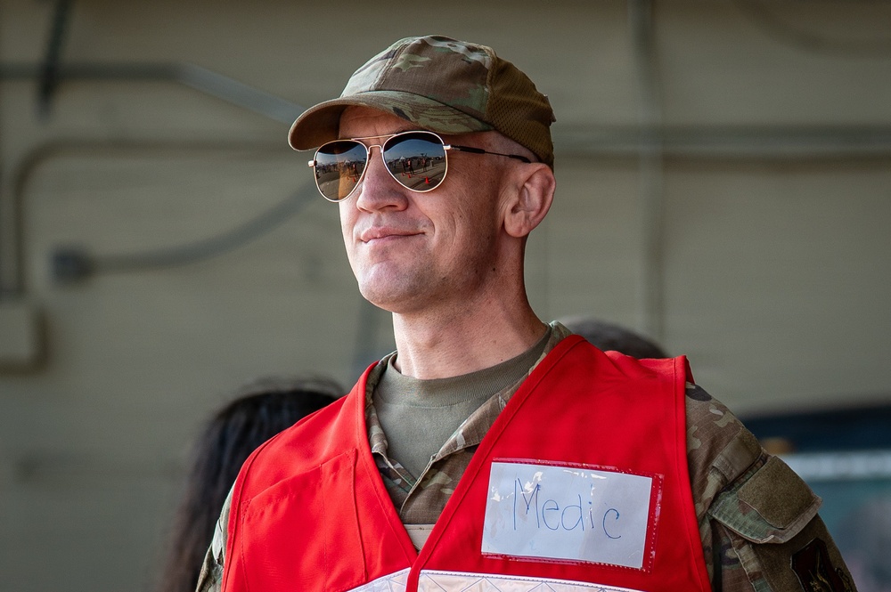 A military medic wearing a red vest with a handwritten 'Medic' label over camouflage uniform is photographed in what appears to be a hangar or military facility. The individual is wearing aviator sunglasses and a multicam/OCP pattern cap, smiling slightly while looking off to the side. The image appears to be taken during a training exercise or operational event.
