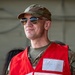 A military medic wearing a red vest with a handwritten 'Medic' label over camouflage uniform is photographed in what appears to be a hangar or military facility. The individual is wearing aviator sunglasses and a multicam/OCP pattern cap, smiling slightly while looking off to the side. The image appears to be taken during a training exercise or operational event.