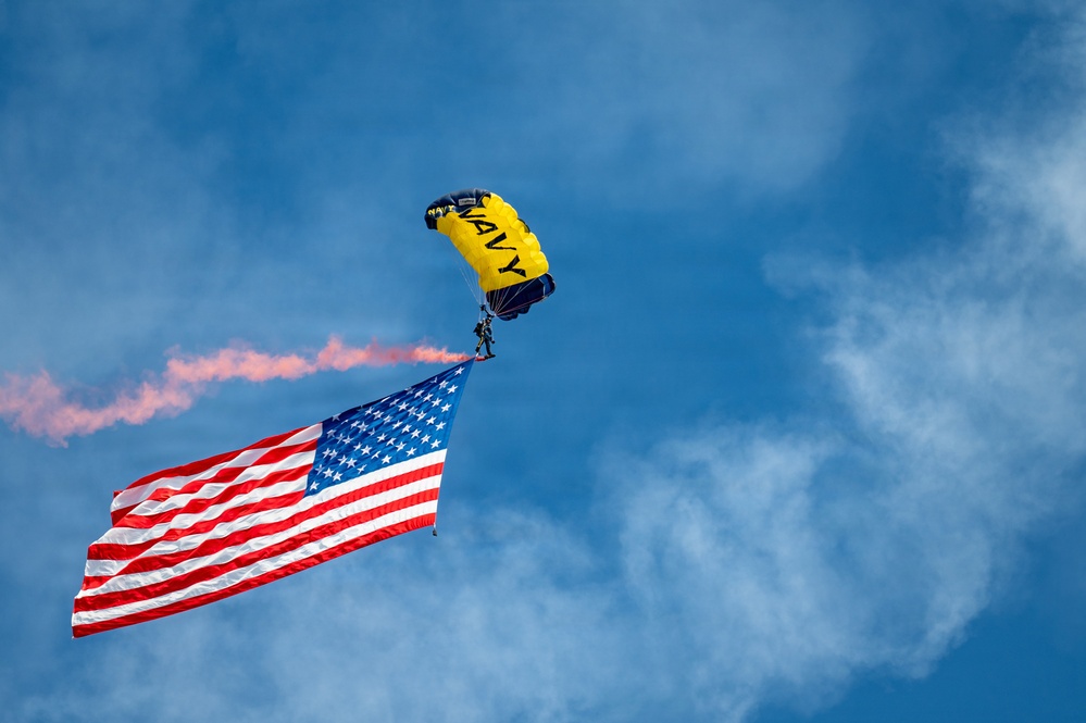 A U.S. Navy parachutist descends under a yellow and navy blue canopy marked 'NAVY' while carrying a large American flag. The jumper is trailing orange/red pyrotechnic smoke from their body, creating a dramatic visual effect against a blue sky with scattered clouds. This appears to be a military demonstration or air show performance.