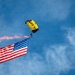 A U.S. Navy parachutist descends under a yellow and navy blue canopy marked 'NAVY' while carrying a large American flag. The jumper is trailing orange/red pyrotechnic smoke from their body, creating a dramatic visual effect against a blue sky with scattered clouds. This appears to be a military demonstration or air show performance.