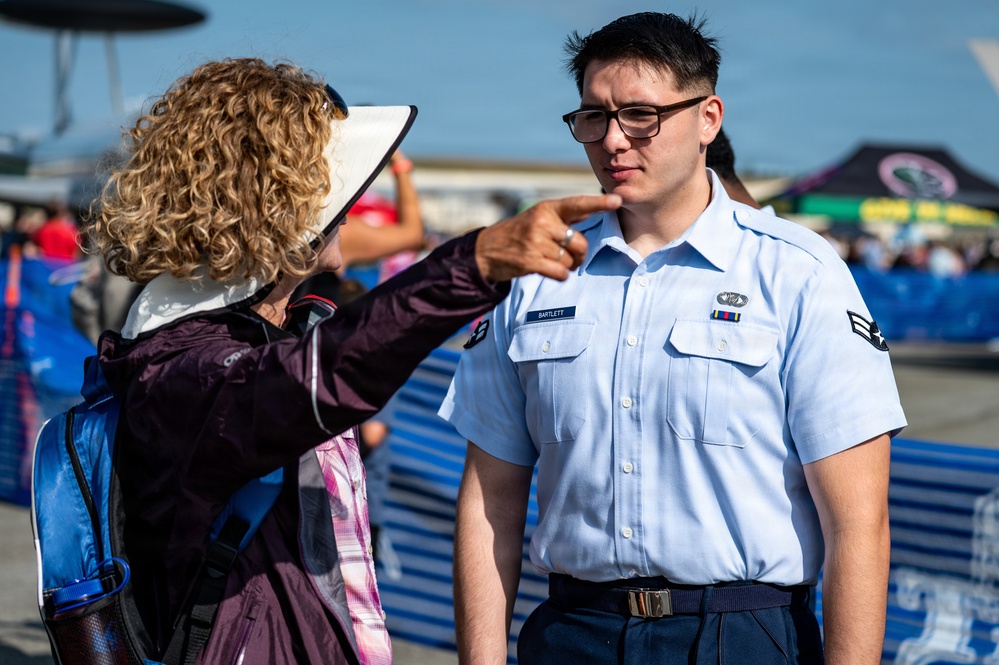 A U.S. Air Force Airman in service dress uniform, identified by the name tape 'BARTLETT,' is being pointed at or directed by a civilian woman wearing a wide-brimmed hat and carrying a backpack. The interaction appears to take place at an outdoor air show event, with aircraft, crowd barriers, and event tents visible in the background. The airman appears attentive or slightly puzzled by the gesture.