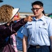 A U.S. Air Force Airman in service dress uniform, identified by the name tape 'BARTLETT,' is being pointed at or directed by a civilian woman wearing a wide-brimmed hat and carrying a backpack. The interaction appears to take place at an outdoor air show event, with aircraft, crowd barriers, and event tents visible in the background. The airman appears attentive or slightly puzzled by the gesture.