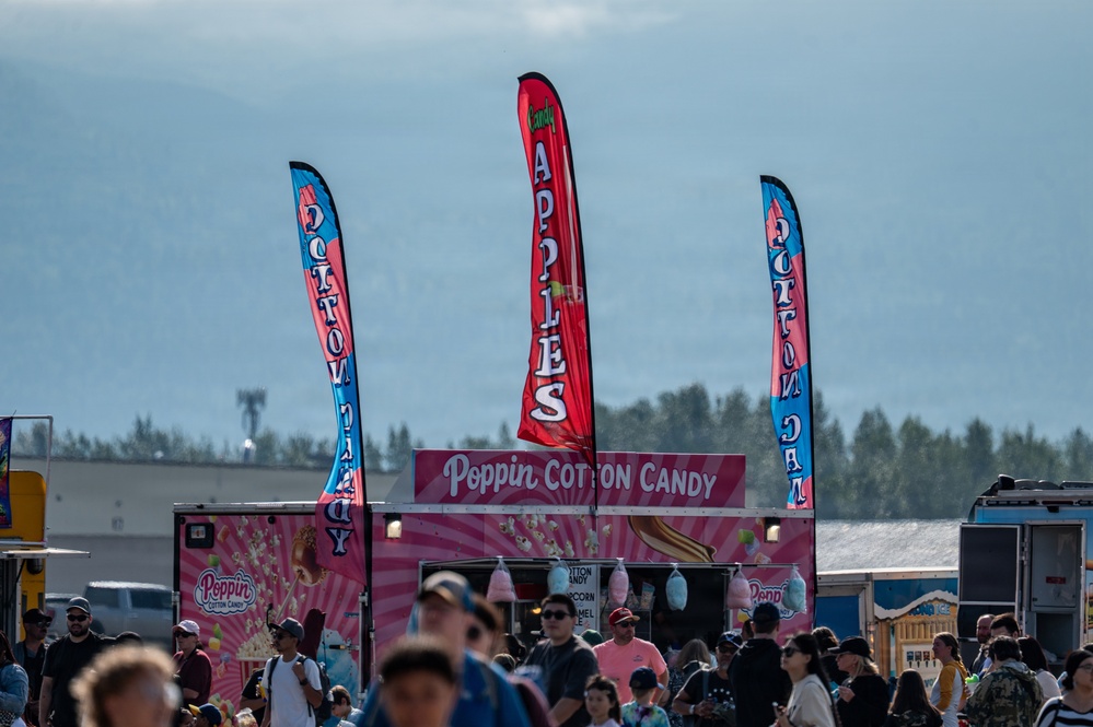A busy outdoor fair or festival scene featuring a 'Poppin Cotton Candy' food vendor truck prominently in the center. Three tall feather/blade banners advertise 'Cotton Candy' and 'Candy Apples' above the crowd. The backdrop includes a treeline and what appears to be mountains or hills under a hazy sky.