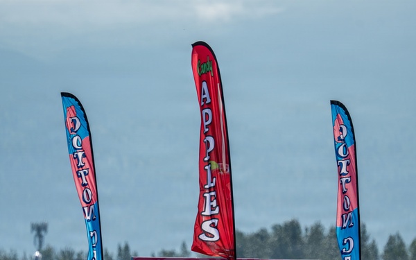 A busy outdoor fair or festival scene featuring a 'Poppin Cotton Candy' food vendor truck prominently in the center. Three tall feather/blade banners advertise 'Cotton Candy' and 'Candy Apples' above the crowd. The backdrop includes a treeline and what appears to be mountains or hills under a hazy sky.