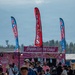 A busy outdoor fair or festival scene featuring a 'Poppin Cotton Candy' food vendor truck prominently in the center. Three tall feather/blade banners advertise 'Cotton Candy' and 'Candy Apples' above the crowd. The backdrop includes a treeline and what appears to be mountains or hills under a hazy sky.