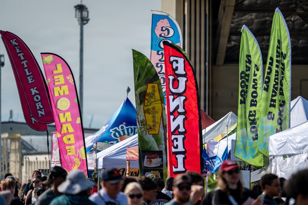 An outdoor food festival or fair with numerous vendor booths marked by colorful feather/blade banners advertising various food and drink items. A large crowd of attendees is visible in the foreground browsing the vendors. The event appears to be held in a large open area, possibly near a stadium or convention center.