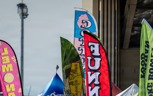 An outdoor food festival or fair with numerous vendor booths marked by colorful feather/blade banners advertising various food and drink items. A large crowd of attendees is visible in the foreground browsing the vendors. The event appears to be held in a large open area, possibly near a stadium or convention center.