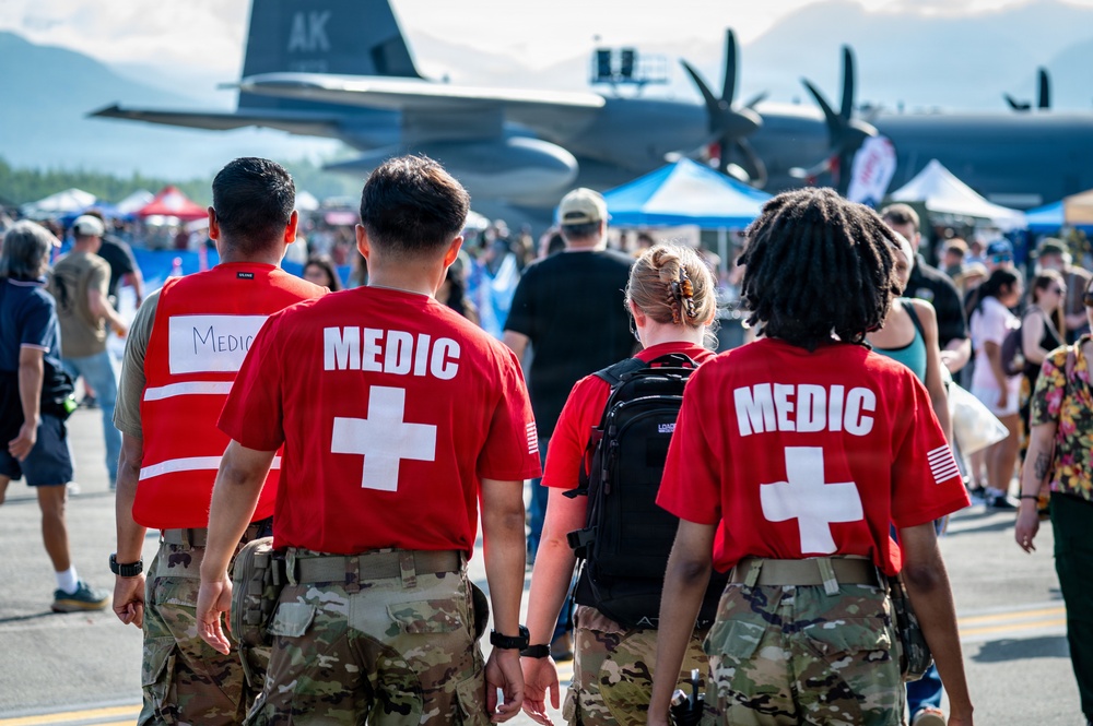Three military medics wearing red 'MEDIC' shirts with white crosses over camouflage pants are walking through a crowded outdoor airshow. A large military transport aircraft, likely a C-130 Hercules, is visible in the background with 'AK' markings, suggesting this is in Alaska. The medics appear to be on patrol duty providing medical coverage for the public event.