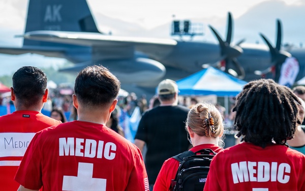 Three military medics wearing red 'MEDIC' shirts with white crosses over camouflage pants are walking through a crowded outdoor airshow. A large military transport aircraft, likely a C-130 Hercules, is visible in the background with 'AK' markings, suggesting this is in Alaska. The medics appear to be on patrol duty providing medical coverage for the public event.
