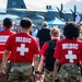 Three military medics wearing red 'MEDIC' shirts with white crosses over camouflage pants are walking through a crowded outdoor airshow. A large military transport aircraft, likely a C-130 Hercules, is visible in the background with 'AK' markings, suggesting this is in Alaska. The medics appear to be on patrol duty providing medical coverage for the public event.