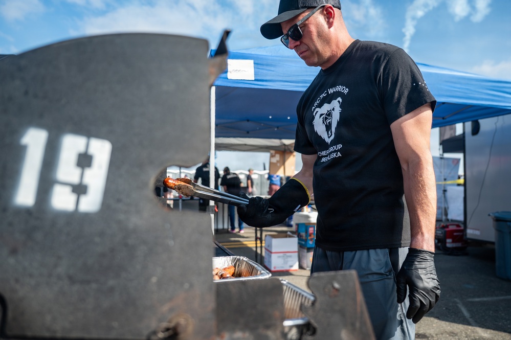 A man wearing a black 'Arctic Warrior' t-shirt and black gloves is grilling sausages at what appears to be an outdoor BBQ competition or community event. He is using tongs to handle meat near a large smoker/grill marked with the number '19'. A blue canopy tent and other participants are visible in the background.