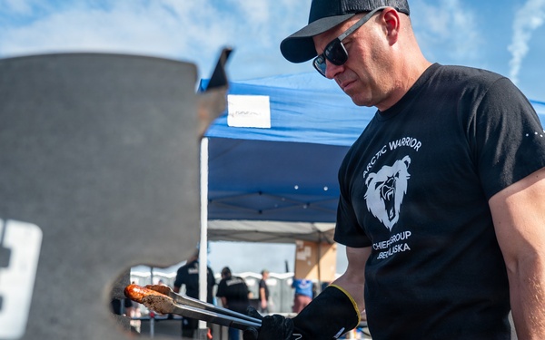 A man wearing a black 'Arctic Warrior' t-shirt and black gloves is grilling sausages at what appears to be an outdoor BBQ competition or community event. He is using tongs to handle meat near a large smoker/grill marked with the number '19'. A blue canopy tent and other participants are visible in the background.