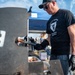 A man wearing a black 'Arctic Warrior' t-shirt and black gloves is grilling sausages at what appears to be an outdoor BBQ competition or community event. He is using tongs to handle meat near a large smoker/grill marked with the number '19'. A blue canopy tent and other participants are visible in the background.