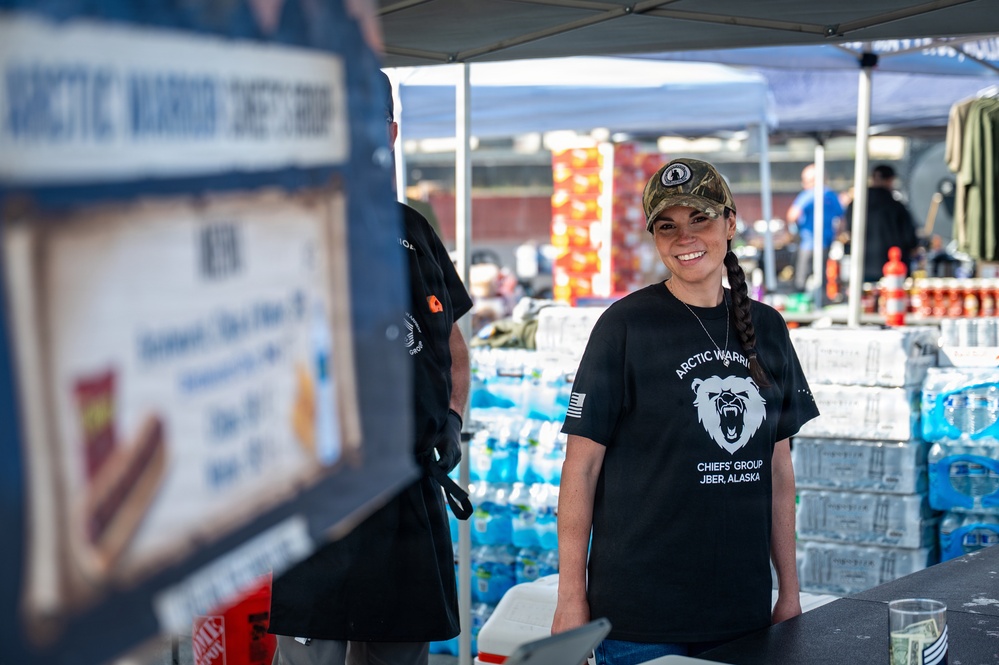 A smiling woman wearing a black 'Arctic Warriors Chiefs' Group JBER, Alaska' t-shirt and camouflage cap stands behind a booth or table at an outdoor event. She appears to be volunteering or working at what looks like a supply distribution or community event, with large quantities of bottled water and other supplies stacked behind her. A second person in a black shirt is partially visible to her left.