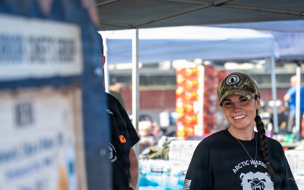 A smiling woman wearing a black 'Arctic Warriors Chiefs' Group JBER, Alaska' t-shirt and camouflage cap stands behind a booth or table at an outdoor event. She appears to be volunteering or working at what looks like a supply distribution or community event, with large quantities of bottled water and other supplies stacked behind her. A second person in a black shirt is partially visible to her left.