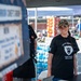 A smiling woman wearing a black 'Arctic Warriors Chiefs' Group JBER, Alaska' t-shirt and camouflage cap stands behind a booth or table at an outdoor event. She appears to be volunteering or working at what looks like a supply distribution or community event, with large quantities of bottled water and other supplies stacked behind her. A second person in a black shirt is partially visible to her left.