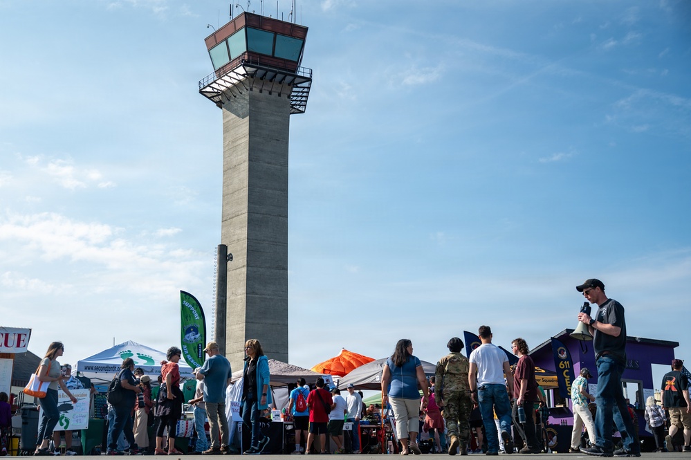 An outdoor public event or air show is taking place on an airport tarmac, with a prominent concrete air traffic control tower dominating the background. Numerous attendees are milling about vendor booths and display tents under a partly cloudy sky. The scene suggests a community open house or aviation-themed festival with multiple organizational exhibitors present.