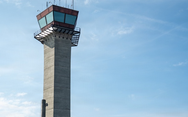 An outdoor public event or air show is taking place on an airport tarmac, with a prominent concrete air traffic control tower dominating the background. Numerous attendees are milling about vendor booths and display tents under a partly cloudy sky. The scene suggests a community open house or aviation-themed festival with multiple organizational exhibitors present.