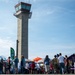 An outdoor public event or air show is taking place on an airport tarmac, with a prominent concrete air traffic control tower dominating the background. Numerous attendees are milling about vendor booths and display tents under a partly cloudy sky. The scene suggests a community open house or aviation-themed festival with multiple organizational exhibitors present.
