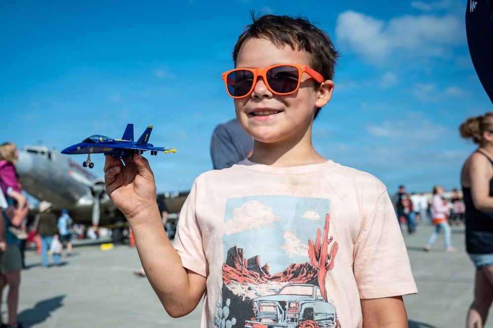 A young boy wearing bright orange sunglasses holds up a Blue Angels F/A-18 toy jet at what appears to be an air show. He is smiling and wearing a light pink t-shirt with a desert landscape graphic. In the background, a vintage military aircraft and a crowd of spectators are visible on an airfield tarmac.