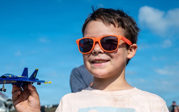 A young boy wearing bright orange sunglasses holds up a Blue Angels F/A-18 toy jet at what appears to be an air show. He is smiling and wearing a light pink t-shirt with a desert landscape graphic. In the background, a vintage military aircraft and a crowd of spectators are visible on an airfield tarmac.