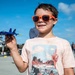A young boy wearing bright orange sunglasses holds up a Blue Angels F/A-18 toy jet at what appears to be an air show. He is smiling and wearing a light pink t-shirt with a desert landscape graphic. In the background, a vintage military aircraft and a crowd of spectators are visible on an airfield tarmac.