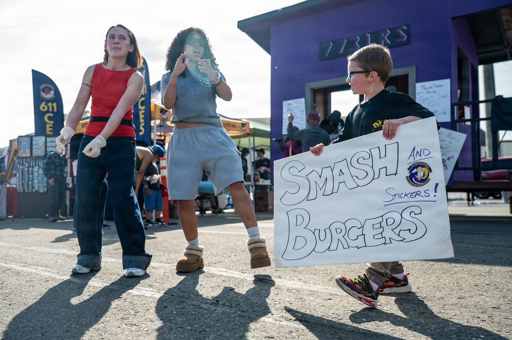 Three young people are at what appears to be an outdoor event or fair near a military base, based on the '611 CE' banner visible in the background. A boy wearing glasses holds a handmade sign reading 'Smash Burgers and Stickers!' while two girls stand nearby, one appearing to dance or move energetically. The setting includes vendor tents, a purple building, and a military squadron banner.