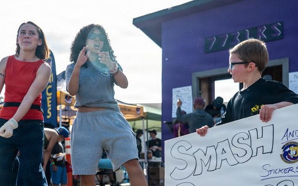 Three young people are at what appears to be an outdoor event or fair near a military base, based on the '611 CE' banner visible in the background. A boy wearing glasses holds a handmade sign reading 'Smash Burgers and Stickers!' while two girls stand nearby, one appearing to dance or move energetically. The setting includes vendor tents, a purple building, and a military squadron banner.