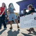 Three young people are at what appears to be an outdoor event or fair near a military base, based on the '611 CE' banner visible in the background. A boy wearing glasses holds a handmade sign reading 'Smash Burgers and Stickers!' while two girls stand nearby, one appearing to dance or move energetically. The setting includes vendor tents, a purple building, and a military squadron banner.